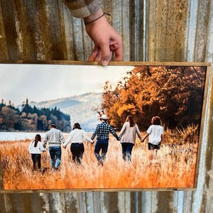 May include: A framed photograph of a family of six walking through a field of tall grass. The family is walking away from the camera, and the background is a forest and a lake. The photo is in a rustic wooden frame.