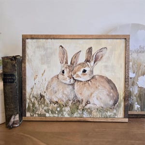 May include: A framed painting depicting two brown rabbits cuddling in a field of green grass. The rabbits have white fur on their bellies and are looking at each other.
