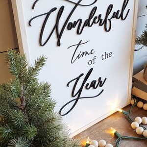 May include: A white wooden sign with the phrase "It's the most Wonderful Time of the Year" in black script. A small evergreen tree in a burlap pot and a string of lights with wooden beads are in the foreground.