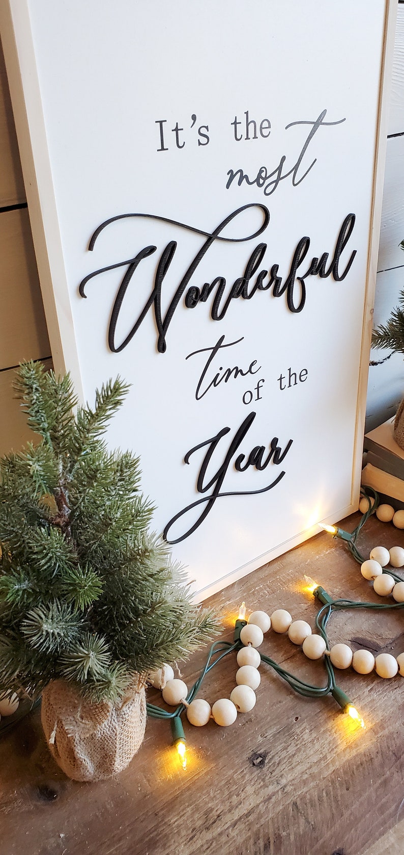May include: A white wooden sign with the phrase "It's the most Wonderful Time of the Year" in black script. A small evergreen tree in a burlap pot and a string of lights with wooden beads are in the foreground.