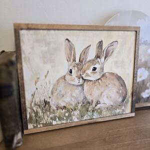 May include: A framed painting depicting two brown rabbits cuddling in a field of green grass. The painting has a rustic, vintage feel.