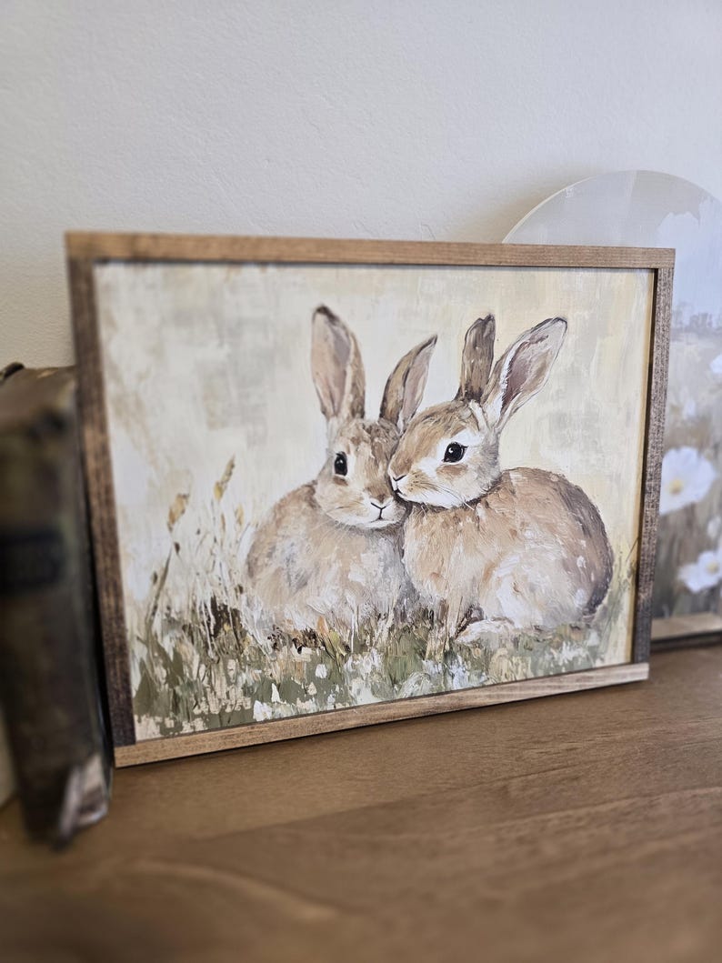 May include: A framed painting depicting two brown rabbits cuddling in a field of green grass. The painting has a rustic, vintage feel.