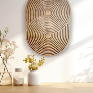 May include: A wooden wall decor with a geometric pattern of concentric circles and ovals in a light brown color. The decor is hanging on a white wall above a wooden countertop with two vases and flowers.