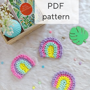 May include: Three handmade crochet rainbow coasters in pastel colors: pink, purple, yellow, and light blue. The image also includes a green leaf cutout, star-shaped confetti, and a book with the text "Rain in the Mountains Ruskin Bond". The text "PDF pattern" is displayed.