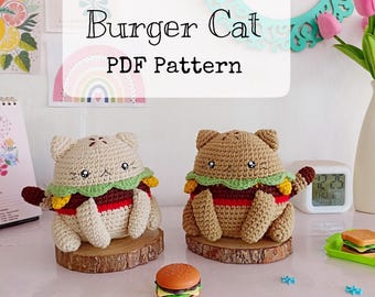 Op de afbeelding: Twee gehaakte kattenknuffels in de vorm van hamburgers, één crème en één beige, met geborduurde gezichtjes, groene sla en rode tomatenlagen. Een klein plastic hamburgerspeeltje staat ertussen, met een bordje waarop Burger Cat PDF Pattern staat.