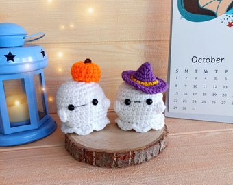 Puede incluir: Dos adornos de fantasmas de crochet con diferentes sombreros. Un fantasma tiene un sombrero de calabaza naranja y el otro un sombrero de bruja morado. Los fantasmas están sentados sobre una base de madera.