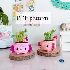 Puede incluir: Dos macetas de axolotl de crochet rosa con plantas verdes. Los axolotls tienen ojos negros y detalles rosas. Las macetas están sobre rodajas de madera. El texto "PDF pattern!" está escrito en letras blancas sobre un fondo rosa.