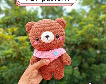 Op de afbeelding: Een bruine gehaakte teddybeer die een roze sjaal draagt met een blauwe bloem. De beer wordt in een hand gehouden tegen een groene, bladerrijke achtergrond. De tekst "PDF pattern" is zichtbaar bovenaan de afbeelding.