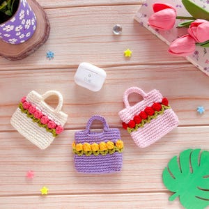 Pode incluir: Três pequenas bolsas de mão de crochê em creme, lavanda e rosa, cada uma adornada com uma fileira de enfeites florais coloridos. Uma capa branca para fones de ouvido sem fio está por perto. As bolsas têm alças pequenas.