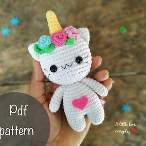 Puede incluir: Un unicornio de crochet blanco con un corazón rosa en el pecho. El unicornio tiene un cuerno amarillo y una corona de flores con flores rosas, azules y verdes. El unicornio está siendo sostenido en una mano.