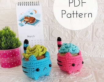 May include: Two crocheted cat planter toys, one blue and one pink, with green succulent plants inside. The cat planters have black stripes and cute faces. The word "PDF Pattern" is written in a circle in the background.