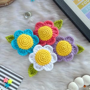 Könnte beinhalten: Vier gehäkelte Blumen in Blau, Rosa, Weiß und Lila, jede mit einem gelben Zentrum und grünen Blättern. Die Blumen sind auf einer weißen, flauschigen Oberfläche angeordnet. Die Blumen sind handgefertigt.