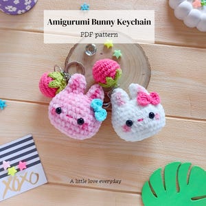 May include: Two crocheted amigurumi bunny keychains, one pink and one white, each featuring black eyes, pink cheeks, and a small bow. The pink bunny has a blue bow, and the white bunny has a pink bow. Both include a small crocheted strawberry charm.