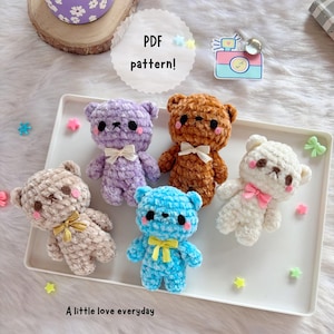 以下が含まれることがあります： ラベンダー、茶色、白、ベージュ、青色の5色の手編みテディベア。各クマには蝶ネクタイとバラ色の頬があります。画像には「PDF pattern！」と「A little love everyday」というテキストが含まれています。