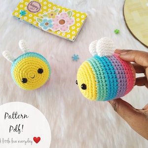 May include: Two crocheted bee plushies. One bee is yellow with blue and white stripes. The other bee is yellow with rainbow stripes. Both bees have black eyes and a smiling mouth.