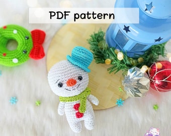 May include: A crocheted snowman amigurumi toy with a blue hat, green scarf, and red buttons. The snowman is sitting on a wooden surface with a blue lantern and Christmas ornaments.