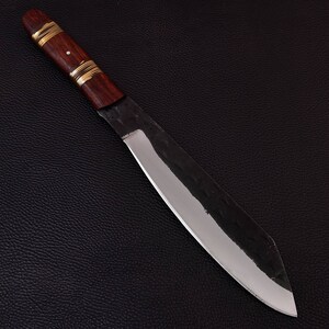 May include: A long, sharp, silver knife with a dark brown wooden handle and gold accents. The knife is lying on a black textured surface.