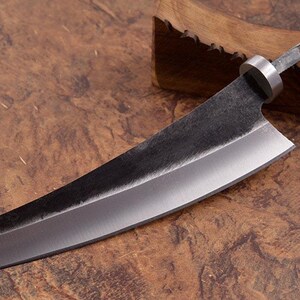 Peut inclure: Un couteau de chef de style japonais avec une lame sombre et texturée et une bague argentée. Le couteau a un bord droit et une pointe carrée. Le manche est une continuation de la soie de la lame. Le couteau est présenté sur un bloc de bois.