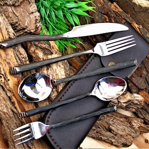 May include: A handcrafted flatware set featuring a knife, fork, and two spoons, with a dark, textured handle design. The set includes a leather carrying case, all displayed on a rustic wooden surface. The knife blade is approximately 9 inches long.