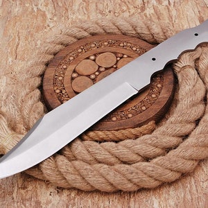 May include: A silver knife blade with a rounded tip and a handle with three holes. The blade is lying on a brown rope and a wooden circular base with a carved design.