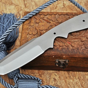 Puede incluir: Una hoja de cuchillo plateada con punta redondeada y un agujero cerca del mango. La hoja está apoyada en una caja de madera con un diseño floral tallado.