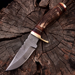 Peut inclure: Un couteau de chasse avec un manche en bois de cerf brun et une lame en acier inoxydable avec un motif Damas. Le couteau est posé sur une surface en bois.