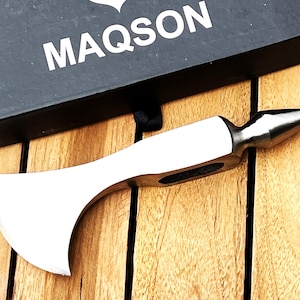May include: A silver-colored axe with a pointed head and a curved blade. The axe is on a wooden surface. The axe is labeled "MAQSON".