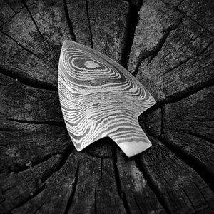 Könnte beinhalten: Ein schwarzer und weißer, gemusterter Metallpfeilspitz mit einer spitzen Spitze, der auf einer rauen, strukturierten Holzoberfläche liegt.