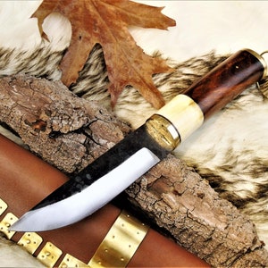 Puede incluir: Un cuchillo con mango negro, mango de madera marrón y pomo de latón. El cuchillo está apoyado en un trozo de corteza y una funda de cuero marrón con detalles en latón.