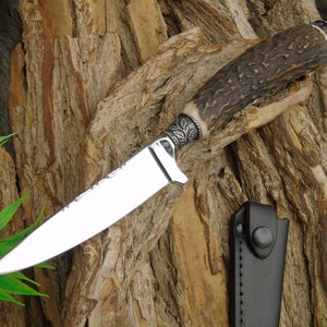 Puede incluir: Un cuchillo de caza con mango de asta de ciervo y hoja de color plateado. El cuchillo tiene una funda de cuero con cierre de presión.