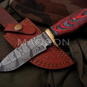 Peut inclure: Un couteau de chasse en acier Damas avec un manche en bois rouge et noir et un fourreau en cuir. Le couteau a un motif ondulé distinctif sur la lame.