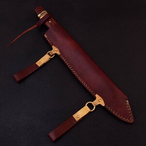 May include: A brown leather sheath with a stitched design and two brass buckles. The sheath has a brown wooden handle sticking out of the top.