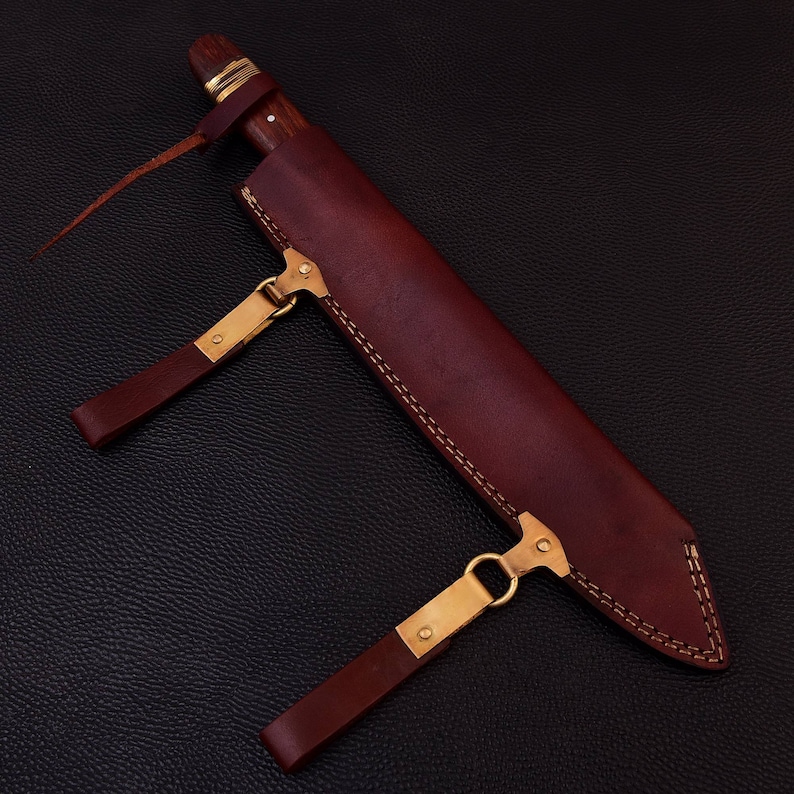 May include: A brown leather sheath with a stitched design and two brass buckles. The sheath has a brown wooden handle sticking out of the top.