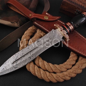 Peut inclure: Un poignard en acier de Damas avec une lame à motifs, un manche noir et des accents décoratifs rouges et dorés. Le poignard repose sur un étui en cuir marron avec une sangle en cuir assortie. Une bobine de corde et un sac camouflage sont en arrière-plan.