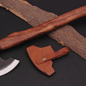 以下が含まれることがあります： 木製の柄と茶色の革製の鞘が付いた小さな黒い斧。斧は湾曲した刃と鋭い刃を持っています。