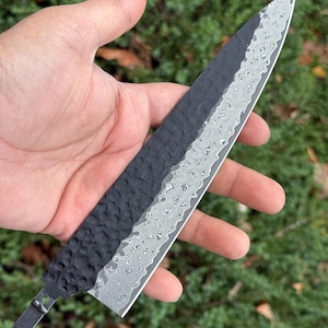May include: A handcrafted chef's knife with a textured black handle and a patterned silver blade. The blade has a unique wave-like design. The knife is held against a backdrop of green foliage.