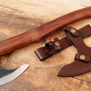 Könnte beinhalten: Eine Axt mit braunem Holzstiel, einer gebogenen Klinge und einer braunen Lederscheide. Die Axt liegt auf einer Holzoberfläche.