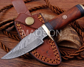 May include: A brown leather sheath with a stitched design holds a small,  damascus steel hunting knife with a wood handle. The handle has a brass accent and a red and gold inlay.