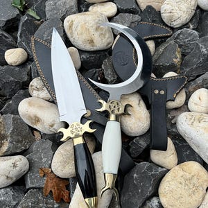 Könnte beinhalten: Zwei verzierte Dolche mit schwarzen und weißen Griffen, goldenen Akzenten und Scheiden. Ein Dolch hat eine gebogene Klinge, der andere eine gerade Klinge. Die Klingen sind silberfarben. Die Dolche werden auf einem Steinhaufen präsentiert.