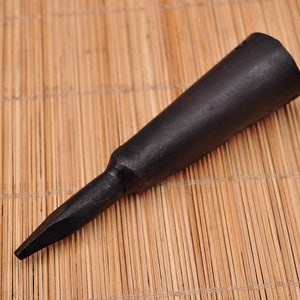 Puede incluir: Una herramienta negra y cónica con un extremo puntiagudo, probablemente utilizada para trabajos de madera. La herramienta está hecha de un material oscuro y descansa sobre una estera de bambú. El diseño presenta un mango cilíndrico que se estrecha hasta un punto afilado. Mide unos 15 cm.