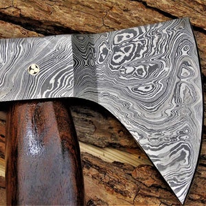 May include: A hand-forged axe with a dark wood handle and a patterned steel head. The head has a unique, swirling pattern of light and dark metal.
