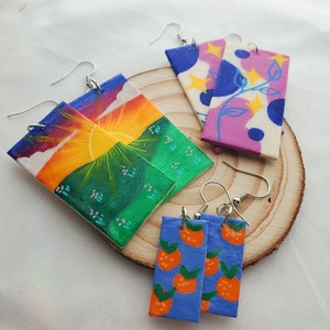 May include: Three pairs of handmade wooden earrings. The first pair features a sunset scene with a green hill, orange sun, and purple clouds. The second pair features a pink and white background with blue circles and yellow stars. The third pair features a blue background with orange oranges.