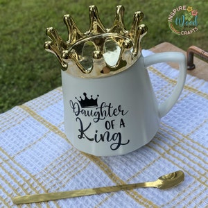May include: White ceramic mug with a gold crown lid and a gold spoon. The mug has the text "Daughter of a King" in black script. The mug is on a white and yellow checkered towel.