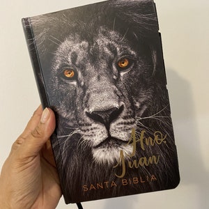 Op de afbeelding: Een hardcover boek met een zwart-wit leeuwenkop op de omslag, met gouden ogen. De tekst "Ano Juan SANTA BIBLIA" is in goud gedrukt. Een zwart lint-bladwijzer is zichtbaar.
