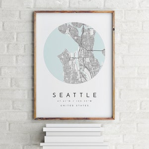 K&ouml;nnte beinhalten: Ein gerahmter Druck einer Karte von Seattle, Washington, mit hellblauem Hintergrund und schwarzen Linien. Der Text "Seattle" ist unter der Karte gedruckt, zusammen mit den Koordinaten "47.61'N / 122.33'W" und "Vereinigte Staaten".