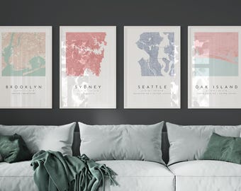 May include: Four framed city maps with a light background and pastel colored city outlines. The maps are labeled "Brooklyn", "Sydney", "Seattle", and "Oak Island". The maps are hanging on a dark gray wall above a white couch with green throw pillows and a green throw blanket.