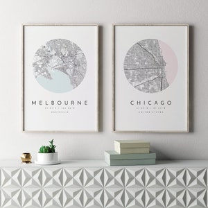 May include: Two framed prints of city maps with a white background. The top print features a black and white map of Melbourne, Australia with a light blue circle around the city. The bottom print features a black and white map of Chicago, United States with a pink circle around the city. The prints are on a white shelf with a small potted succulent plant and two boxes.