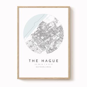 Könnte beinhalten: Ein gerahmter Druck mit einer detaillierten Karte von Den Haag, Niederlande, in einem kreisförmigen Design. Die Karte ist in Schwarzweiß gehalten, mit einem hellblauen Akzent. Der Text "THE HAGUE" und Koordinaten sind darunter.