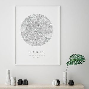 May include: A black and white map print of Paris, France. The map is in a circle with the city name and coordinates below. The print is framed in a white frame and is hanging on a white wall.