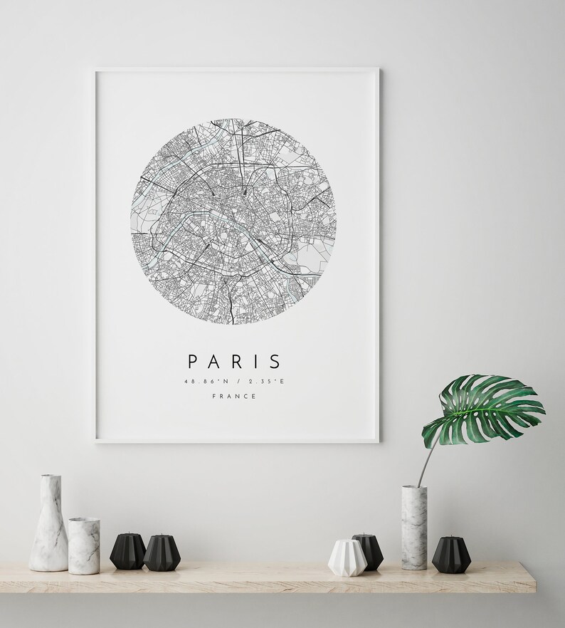 May include: A black and white map print of Paris, France. The map is in a circle with the city name and coordinates below. The print is framed in a white frame and is hanging on a white wall.